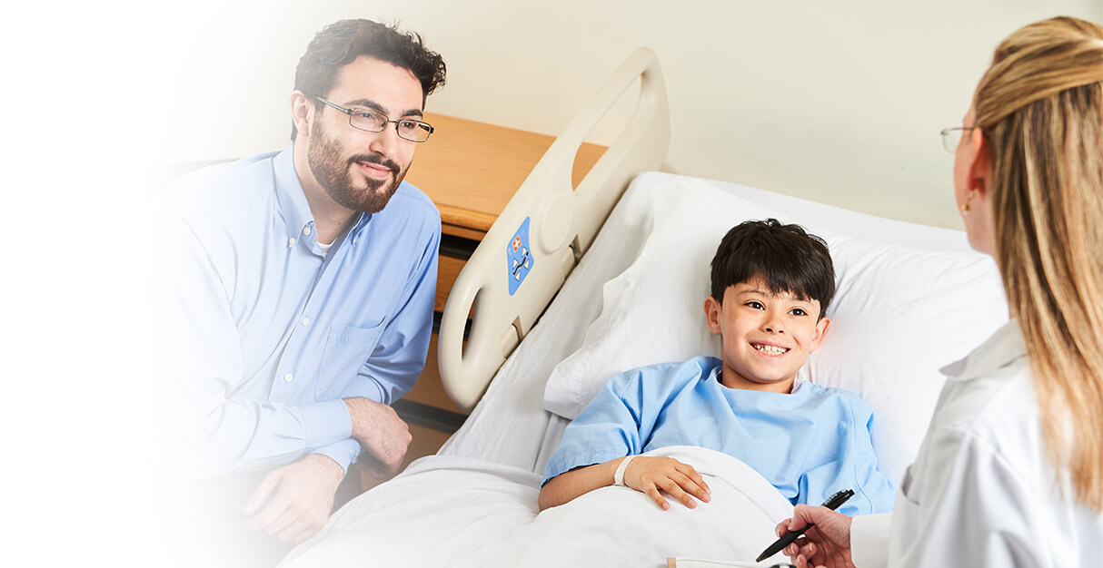 A young boy in a hospital bed talking to a doctor while his father watches.